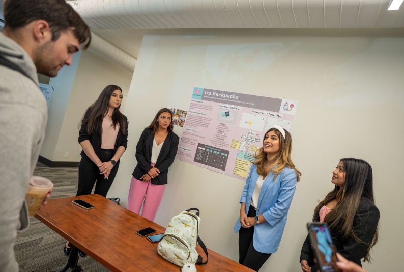 Students stand next to a poster with a backpack on a table in front of them